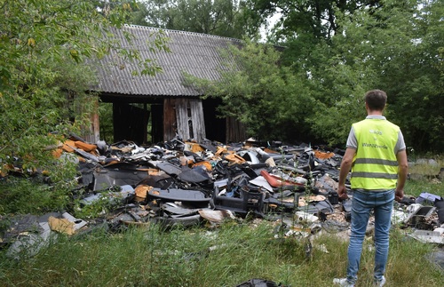 Inspektor Wojewódzkiego Inspektoratu Ochrony Środowiska w Warszawie stoi przed rozrzuconymi na ziemi odpadami z demontażu pojazdów. W tle znajduje się budynek gospodarczy.