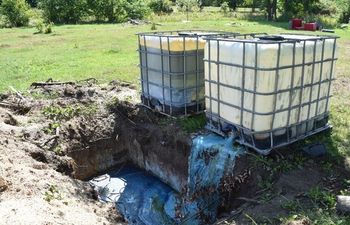 Dwa zbiorniki o pojemności 1000 l stoją na otwartym terenie. Pry nich znajduje się wykop wypełniony niebieską substancją wprowadzoną do gruntu z tych zbiorników.